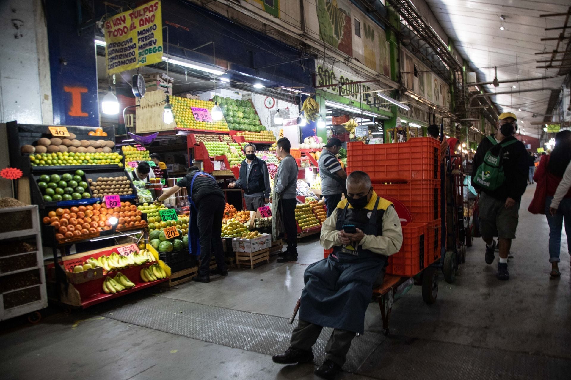 Un diablero espera mirando su celular a un costado de un puesto de frutas en la Central de Abastos